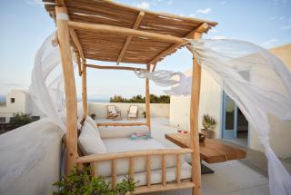 Wooden canopy bed on a terrace with white curtains blowing in the wind two chairs and a small table with two glasses of wine are in the background white buildings and a clear sky are visible hu13788127133455560704.jpg